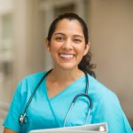 Smiling female healthcare professional looks at the camera while in hospital hallway. She is standing with her arms crossed.